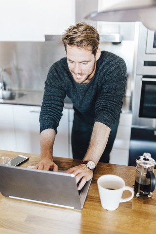 Lächelnder Mann steht in der Küche und benutzt einen Laptop, lizenzfreies Stockfoto