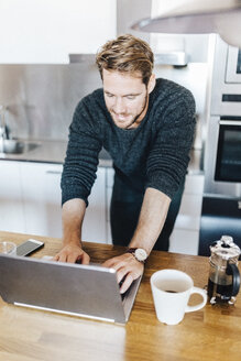 Lächelnder Mann steht in der Küche und benutzt einen Laptop - GIOF03178