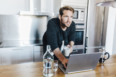 Porträt eines Mannes mit Kaffeetasse und Laptop in der Küche stehend - GIOF03179