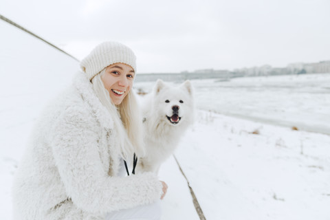 Porträt einer weiß gekleideten lächelnden jungen Frau mit ihrem weißen Hund im Schnee, lizenzfreies Stockfoto