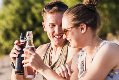 Glückliches junges Paar stößt im Freien mit Flaschen an - UUF11529