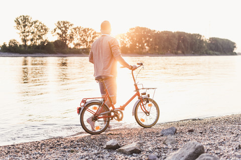 Junger Mann zu Fuß mit Fahrrad am Flussufer bei Sonnenuntergang, lizenzfreies Stockfoto