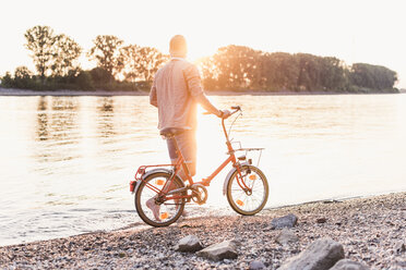 Junger Mann zu Fuß mit Fahrrad am Flussufer bei Sonnenuntergang - UUF11553