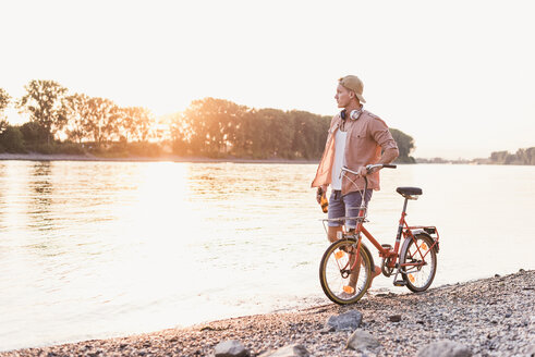 Young man walking with bicycle at the riverbank at sunset - UUF11554