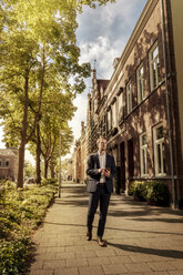 Netherlands, Venlo, businessman with cell phone walking on pavement - KNSF02413