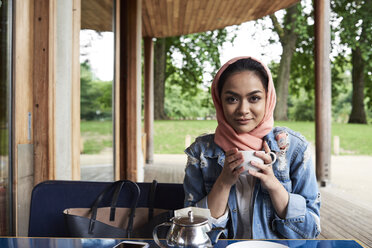 Junge Frau mit Hidschab, die eine Tasse auf der Terrasse eines Cafés hält - IGGF00133