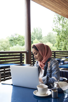 Junge Frau mit Hidschab, die mit ihrem Laptop auf der Terrasse eines Cafés lernt - IGGF00135