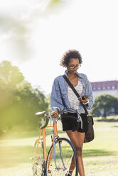 Junge Frau mit Mobiltelefon schiebt Fahrrad im Park - UUF11600