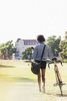 Junge Frau mit Mobiltelefon schiebt Fahrrad im Park - UUF11603