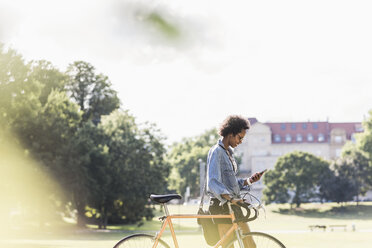 Junge Frau mit Mobiltelefon schiebt Fahrrad im Park - UUF11605