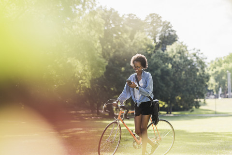 Junge Frau mit Mobiltelefon schiebt Fahrrad im Park, lizenzfreies Stockfoto