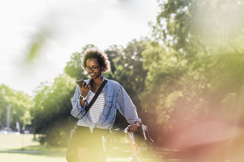 Junge Frau mit Mobiltelefon schiebt Fahrrad im Park, lizenzfreies Stockfoto