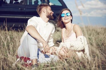 Happy young couple sitting on field next to car - ABIF00018