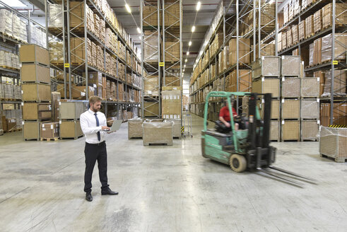 Businessman with laptop in warehouse and worker on forklift - LYF00764