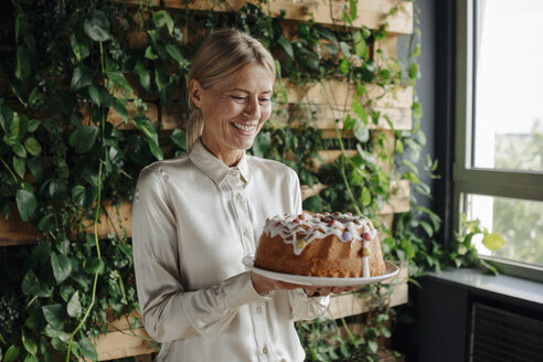 Smiling businesswoman holding birthday cake in green office - JOSF01367