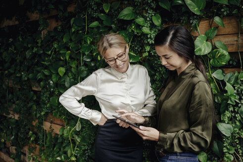 Zwei Geschäftsfrauen in einem grünen Büro mit Blick auf ein Tablet - JOSF01383