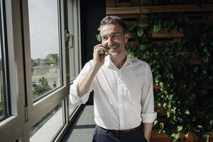 Lächelnder Geschäftsmann mit Mobiltelefon am Fenster - JOSF01392