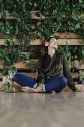 Young woman sitting on floor at wall with climbing plants listening to music - JOSF01408
