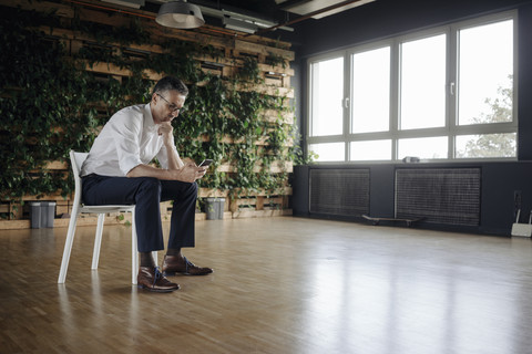 Geschäftsmann sitzt auf einem Stuhl in einem grünen Büro und schaut auf sein Handy, lizenzfreies Stockfoto