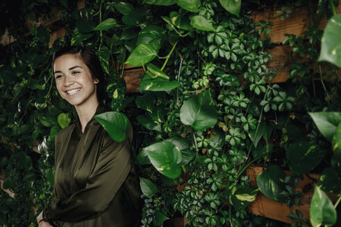 Happy young woman at wall with climbing plants - JOSF01448