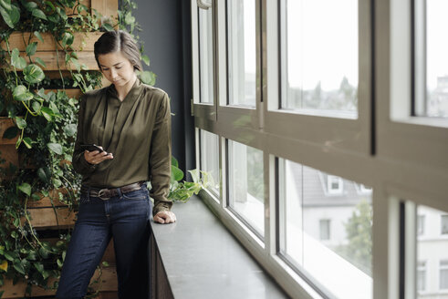 Junge Frau im Büro am Fenster, die auf ihr Mobiltelefon schaut - JOSF01451