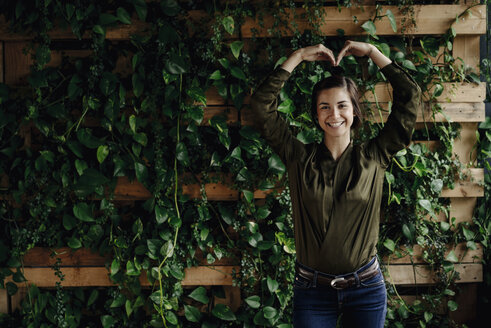 Portait of smiling young woman shaping heart at wall with climbing plants - JOSF01452