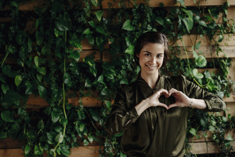 Portait of smiling young woman shaping heart at wall with climbing plants stock photo