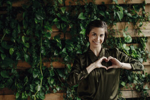 Porträt einer lächelnden jungen Frau, die ein Herz an einer Wand mit Kletterpflanzen formt - JOSF01453