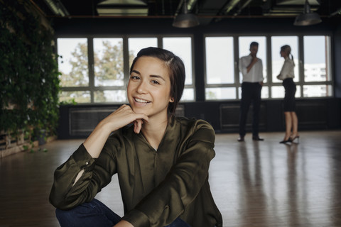 Lächelnde Frau sitzt auf dem Boden im Büro mit zwei Menschen im Hintergrund, lizenzfreies Stockfoto
