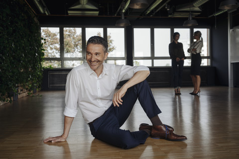 Smiling businessman sitting on the floor in office with two women in background stock photo