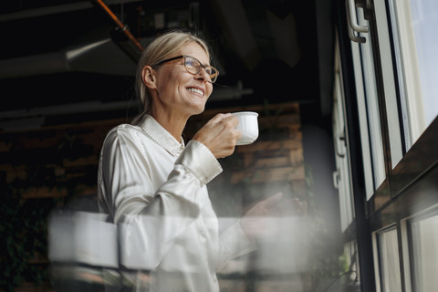 Lächelnde Geschäftsfrau, die eine Tasse Kaffee hält und aus dem Fenster schaut, lizenzfreies Stockfoto