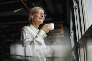 Smiling businesswomanholding cup of coffee looking out of window - JOSF01503