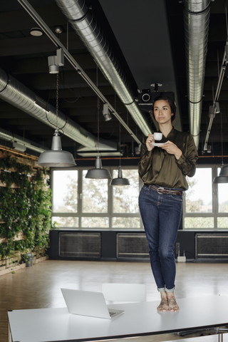 Junge Frau im Büro stehend auf einem Tisch mit einer Tasse Kaffee, lizenzfreies Stockfoto