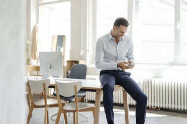 Lächelnder Geschäftsmann im Büro, der auf sein Mobiltelefon schaut - KNSF02418