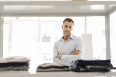 Portrait of confident businessman standing at shelf with clothes - KNSF02423