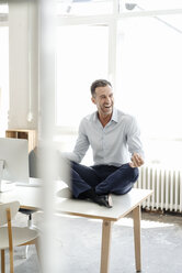 Happy businessman sitting on table in office practising yoga - KNSF02437