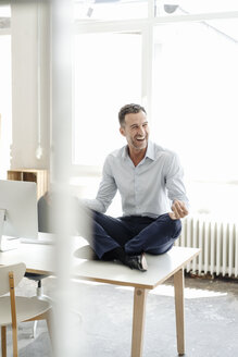 Glücklicher Geschäftsmann sitzt auf einem Tisch im Büro und praktiziert Yoga - KNSF02437