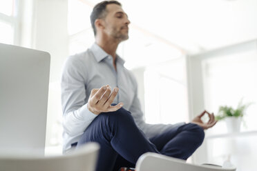 Businessman sitting on table in office practising yoga - KNSF02439