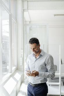 Lächelnder Geschäftsmann im Büro am Fenster, der auf sein Handy schaut - KNSF02450