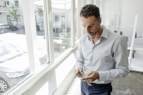 Geschäftsmann im Büro am Fenster und schaut auf sein Handy, lizenzfreies Stockfoto