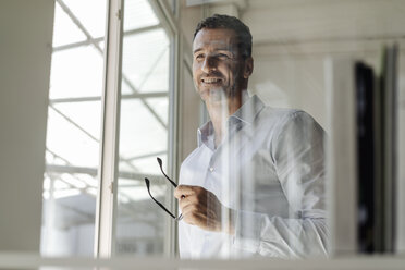 Smiling businessman holding glasses standing at the window - KNSF02457