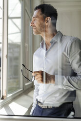 Businessman holding glasses looking out of window - KNSF02458