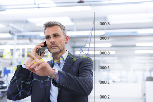 Geschäftsmann mit Handy, der ein Diagramm auf einer Glasscheibe in einer Fabrikhalle betrachtet - DIGF02776
