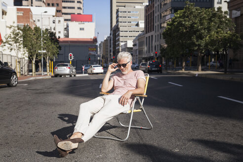 Mature man sitting on chair in the street, wearing sunglasses - WESTF23572