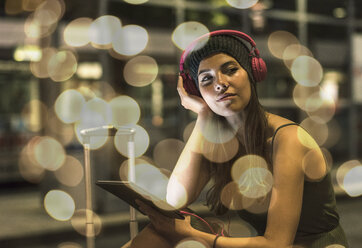 Portrait of young woman with headphones and tablet waiting at station by night - UUF11622