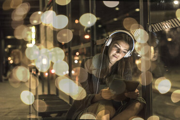 Porträt einer jungen Frau mit Kopfhörern und Tablet, die am nächtlichen Bahnhof wartet - UUF11627