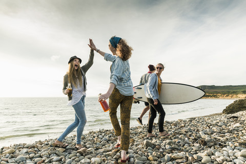 Excited friends with surfboard and drinks on stony beach stock photo