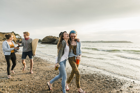 Happy friends with surfboard and drinks walking on stony beach stock photo