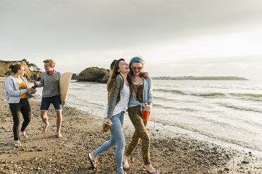 Fröhliche Freunde mit Surfbrett und Getränken spazieren am steinigen Strand - UUF11653