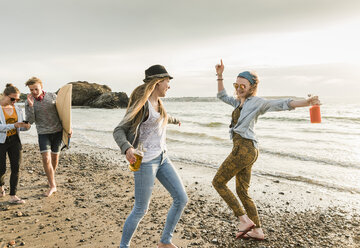 Fröhliche Freunde mit Surfbrett und Getränken spazieren am steinigen Strand - UUF11654